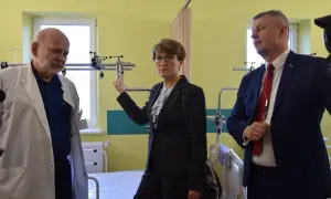 Trzy osoby w sali szpitalnej, dwie w garniturach, jedna w kitlu lekarskim.
