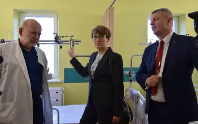 Trzy osoby w sali szpitalnej, dwie w garniturach, jedna w kitlu lekarskim.