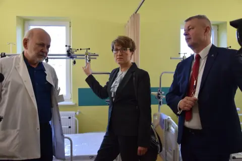 Trzy osoby w sali szpitalnej, dwie w garniturach, jedna w kitlu lekarskim.