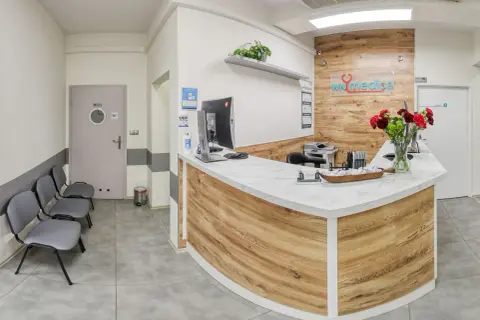 Recepcja kliniki medycznej z ladą, komputerem, krzesłami i bukietem kwiatów.