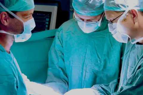 Trzech chirurgów w maseczkach i czepkach operacyjnych pochylonych nad pacjentem.