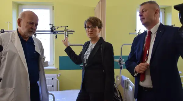 Trzy osoby w sali szpitalnej, dwie w garniturach, jedna w kitlu lekarskim.
