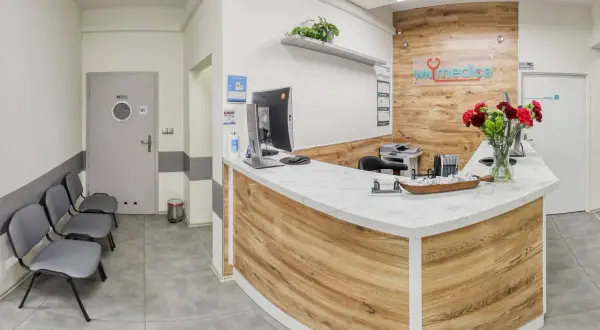 Recepcja kliniki medycznej z ladą, komputerem, krzesłami i bukietem kwiatów.