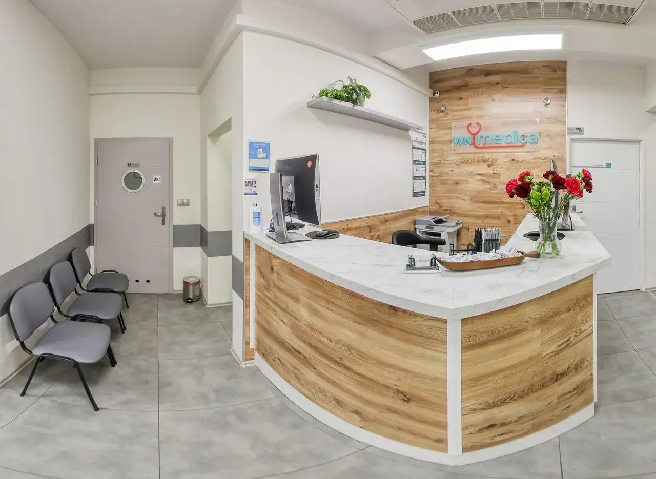 Recepcja kliniki medycznej z ladą, komputerem, krzesłami i bukietem kwiatów.